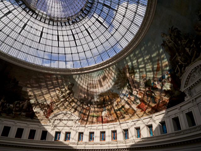 A large glass dome ceiling with intricate metal framework, casting shadows on a mural below depicting historical and mythical scenes.