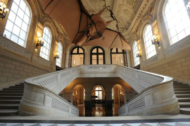 Grand staircase in a historic building with large windows, arched doorways, and a flying machine suspended from the ceiling.