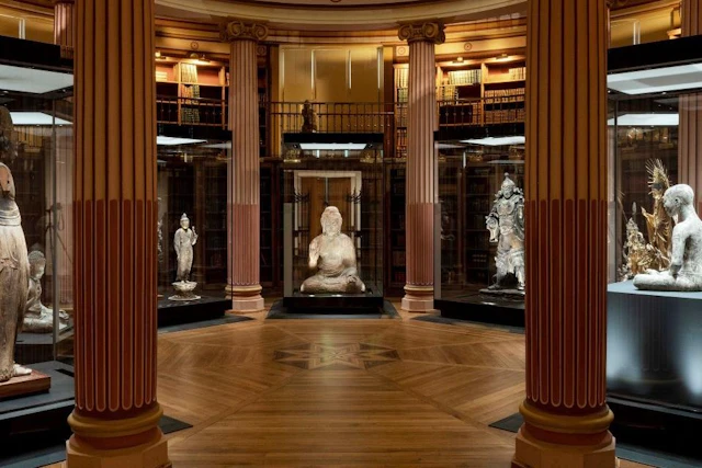 A museum hall with three sculptures displayed in glass cases, surrounded by bookshelves and decorated columns.