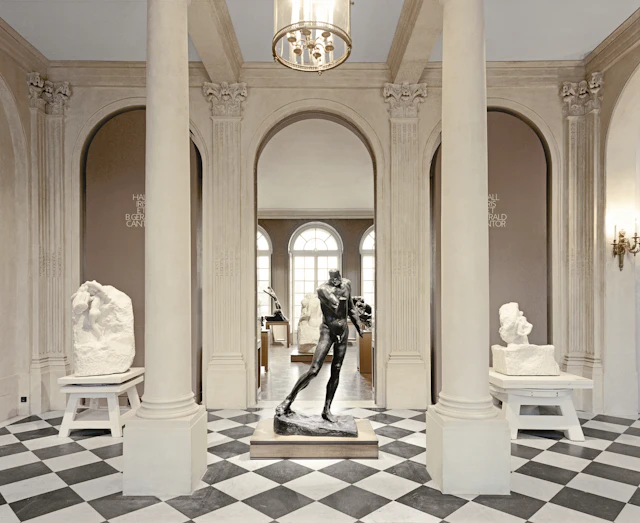 Marble sculptures and a bronze statue in a museum gallery with arched doorways, two columns, checkered floor, and chandeliers.