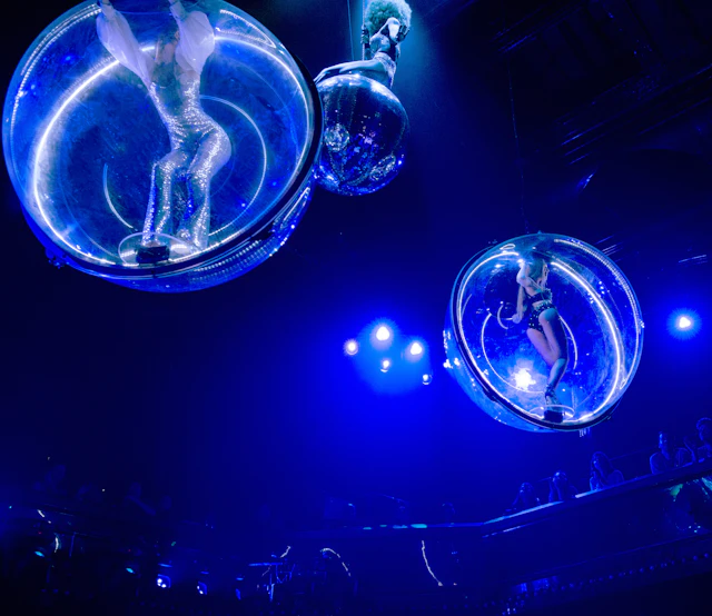Performers in glowing costumes are inside transparent spheres suspended above a stage with blue lighting. Audience visible below.