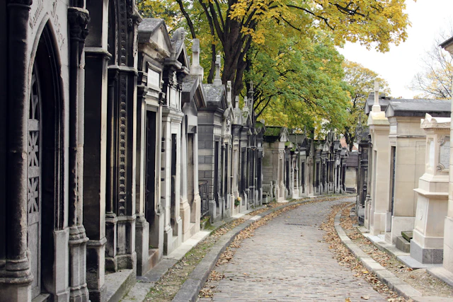 A cobblestone path lined with mausoleums on each side, shaded by green and yellow-leaved trees.